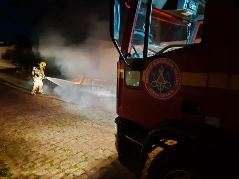 Bombeiro apagando incêndio em veículo durante a noite, com fumaça visível e caminhão dos bombeiros em primeiro plano.