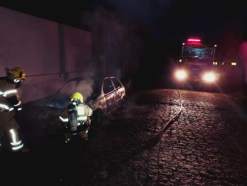 Bombeiros combatendo o incêndio em um carro parcialmente queimado em uma rua estreita, com um caminhão de bombeiros ao fundo.