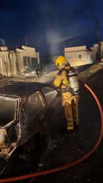 Bombeiro usando mangueira para combater incêndio em carro queimado em uma rua residencial.