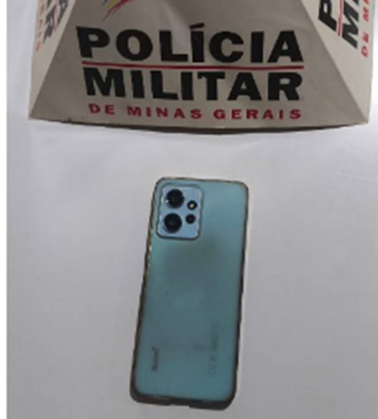 Um celular Xiaomi Redmi Note 12 de cor azul posicionado em primeiro plano, com uma caixa de fundo da Polícia Militar de Minas Gerais visível.