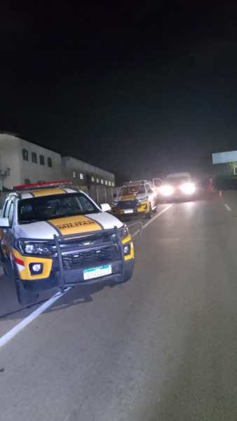 Veículos da Polícia Militar parados à beira da rodovia à noite, em uma operação de fiscalização.