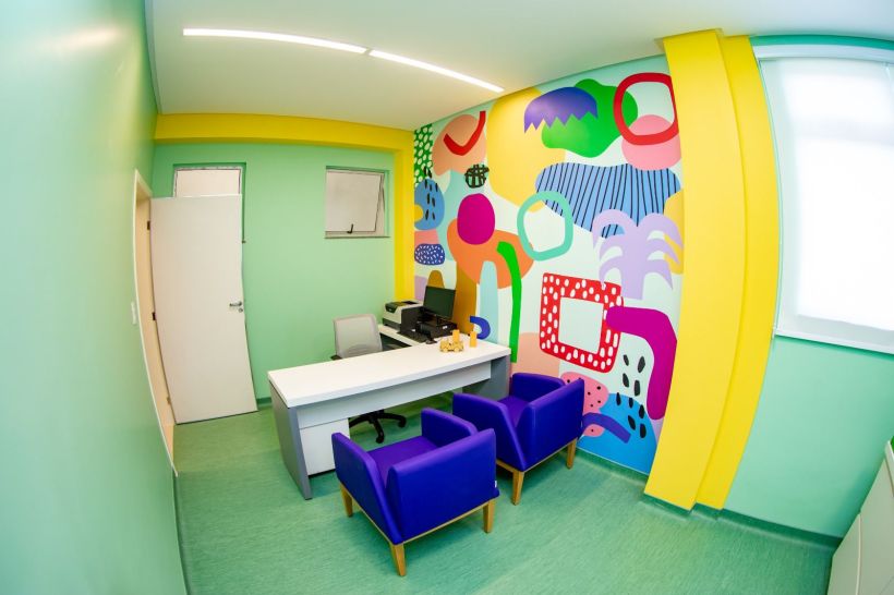 Interior de uma sala de oncopediatria com paredes coloridas e decoração alegre. A sala possui uma mesa de escritório e cadeiras roxas.