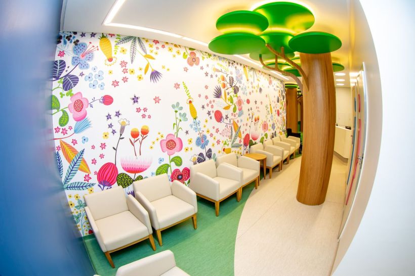 Interior da ala de Oncopediatria, com um mural colorido de flores e pássaros, cadeiras brancas e uma árvore decorativa.