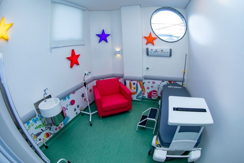 Interior colorido da ala de Oncopediatria, com uma cadeira vermelha, decoração infantil nas paredes e equipamentos médicos.