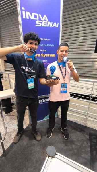 Dois estudantes segurando troféus e medalhas em frente a um painel do Inova SENAI.