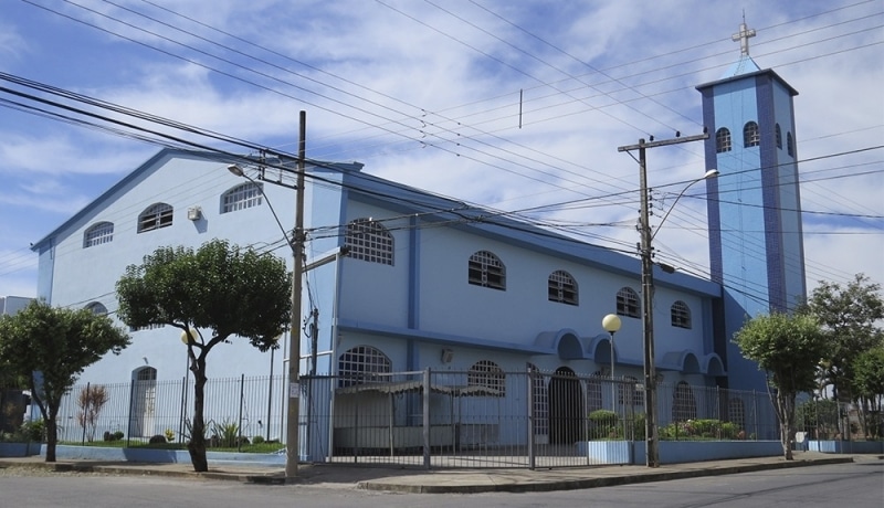 Igreja Nossa Senhora Aparecida em Divinópolis, com fachada azul e torre com cruz, cercada por árvores e postes de luz.