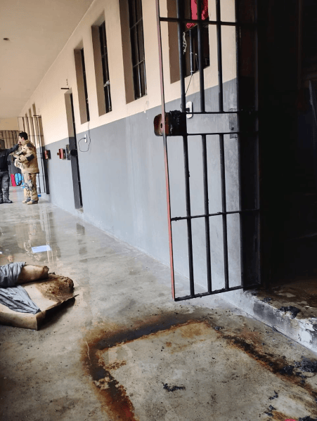 Corredor da Unidade Prisional Juca do Anibal com um portão de cela aberto, chão molhado e sinais de um incêndio recente, sem detentos visíveis.