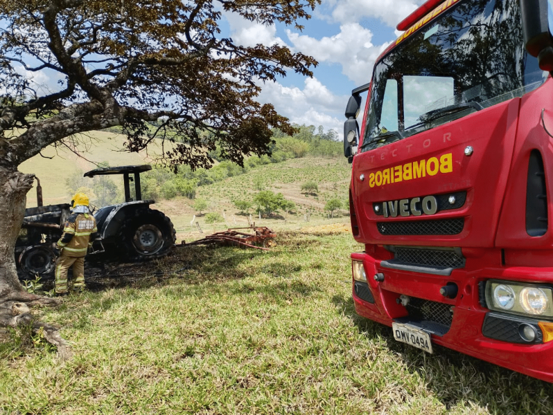 Um caminhão do Corpo de Bombeiros vermelho estacionado em primeiro plano, com um trator queimado ao fundo em uma área rural rodeada por pastagens e árvores.