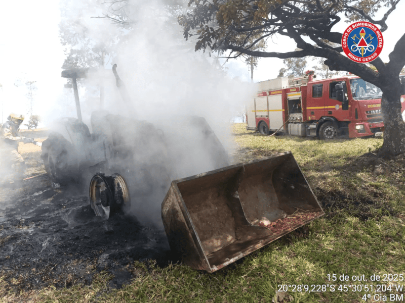 Trator do tipo pá-carregadeira em chamas na zona rural, com fumaça saindo do maquinário; veículos dos bombeiros ao fundo.