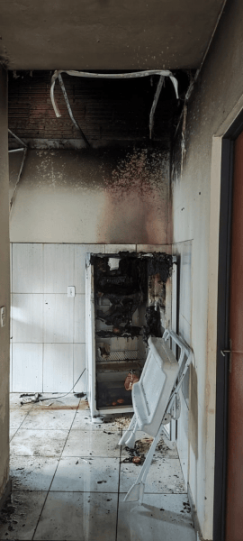 Interior de uma residência com danos significativos causados por incêndio, incluindo uma geladeira queimada e forro de PVC danificado.