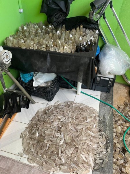 Uma pilha de cristais de quartzo empilhados em um local com paredes verdes, ao lado de uma caixa com cristais, ferramentas e sacos plásticos.