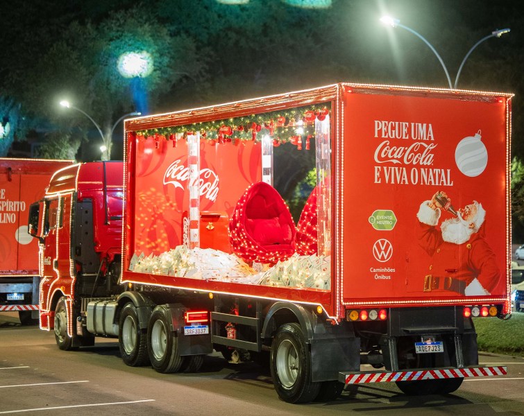 Caminhão decorado da Coca-Cola com luzes de Natal, exibindo um layout festivo e a mensagem 'Pegue uma Coca-Cola e viva o Natal'.