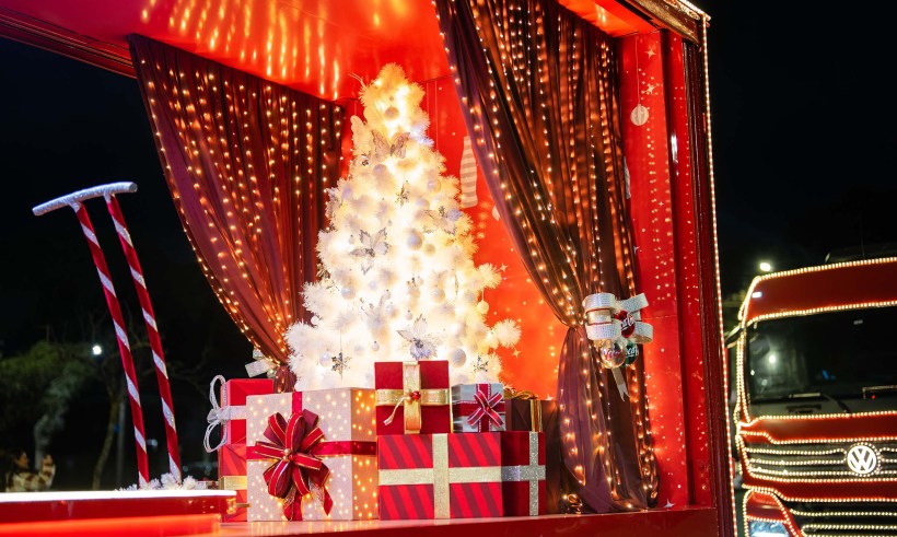 Decoração natalina com uma árvore branca iluminada, presente decorados com fitas, e elementos festivos em um caminhão iluminado ao fundo.