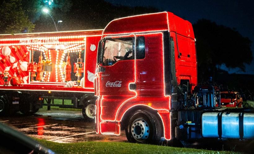 Caminhão iluminado da Coca-Cola com decoração natalina, parado sob a chuva à noite.