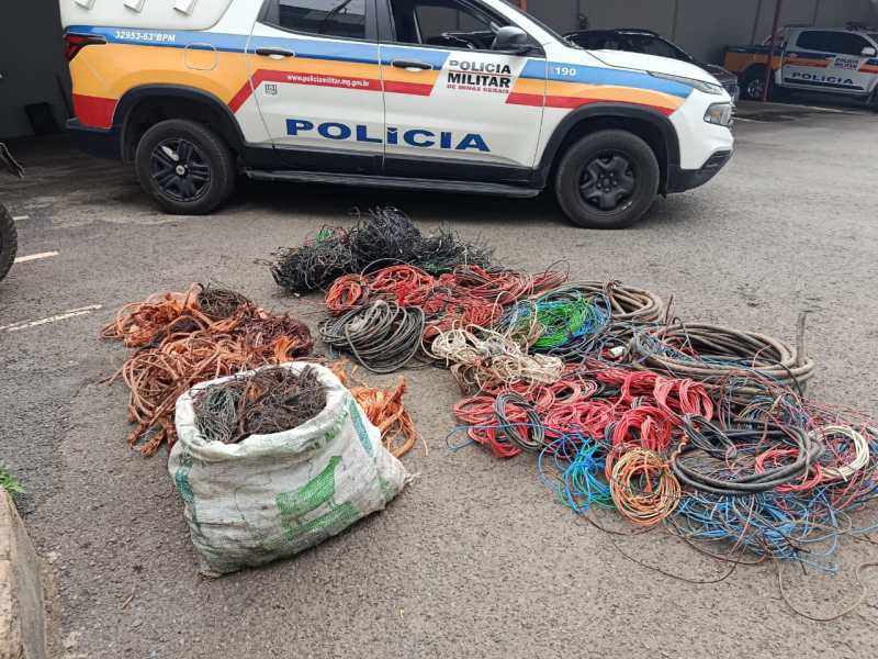 Diversos fios de cobre e cabos emaranhados estão empilhados no chão, ao lado de um veículo da Polícia Militar, que está estacionado em uma área coberta.