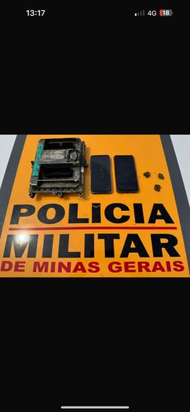 Central automotiva com numeração picotada, dois smartphones e porções de droga sobre placa da Polícia Militar de Minas Gerais.