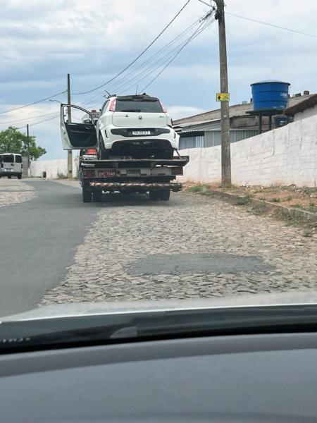 Uma caminhonete de guincho transporta um carro branco com danos significativos na parte dianteira, em uma rua de paralelepípedo. Ao fundo, há postes de energia elétrica e uma casa com muros brancos.