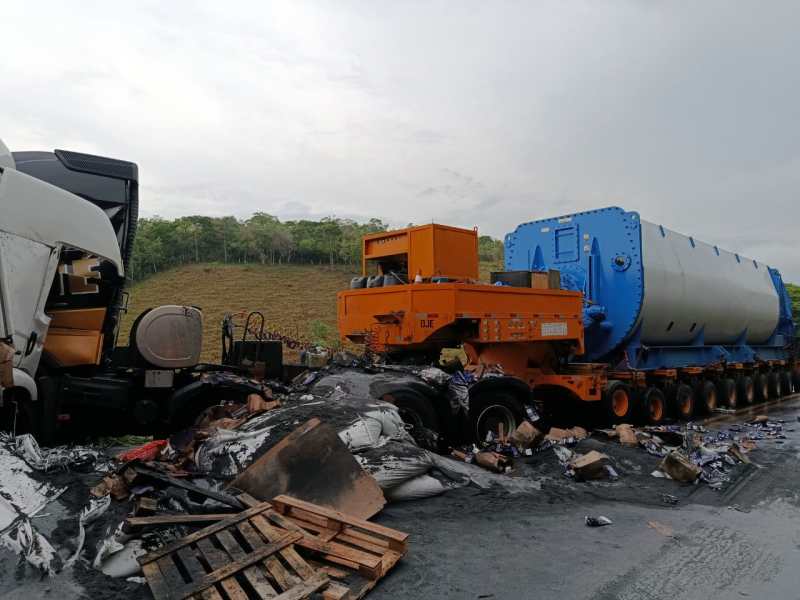 Cenário de colisão entre um caminhão baú e uma carreta de grande porte, com destroços espalhados e carga danificada na pista.