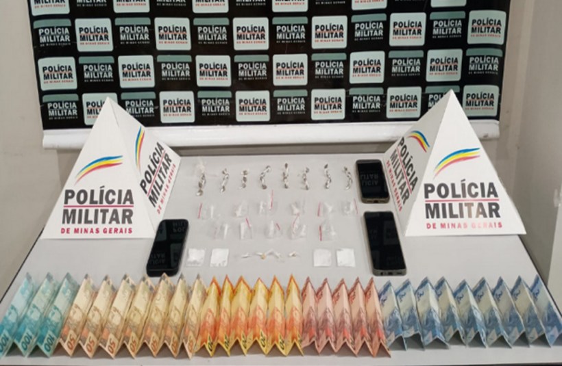 Materiais apreendidos durante operação policial, incluindo drogas, dinheiro e celulares, dispostos sobre uma mesa com sinalização da Polícia Militar de Minas Gerais ao fundo.