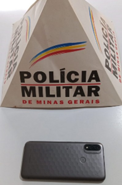 Um celular preto em frente a uma base da Polícia Militar de Minas Gerais, destacando a logo da corporação.
