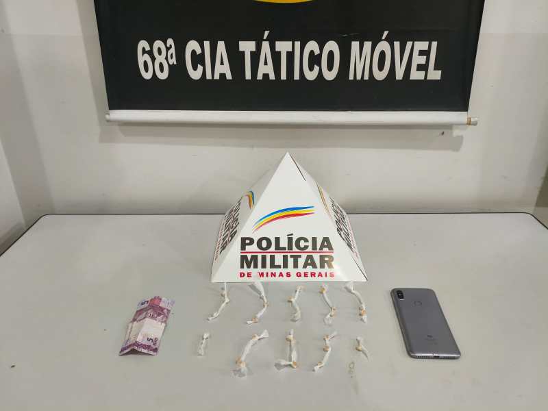Materiais apreendidos pela Polícia Militar de Minas Gerais, incluindo entorpecentes e um celular, exibidos sobre uma mesa com um banner da 68ª Companhia Tático Móvel ao fundo.