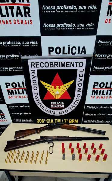 Duas armas de fogo e munições apreendidas pela Polícia Militar de Minas Gerais, com um cartaz informativo ao fundo.