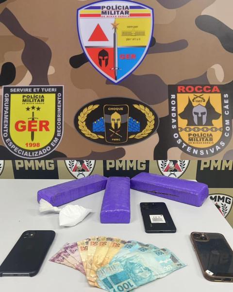 Imagem de uma mesa com drogas, dinheiro e celulares. Abaixo, há três barras roxas que aparentam ser drogas, uma quantidade de dinheiro em notas e dois celulares. Ao fundo, em destaque, estão emblemas da Polícia Militar de Minas Gerais.