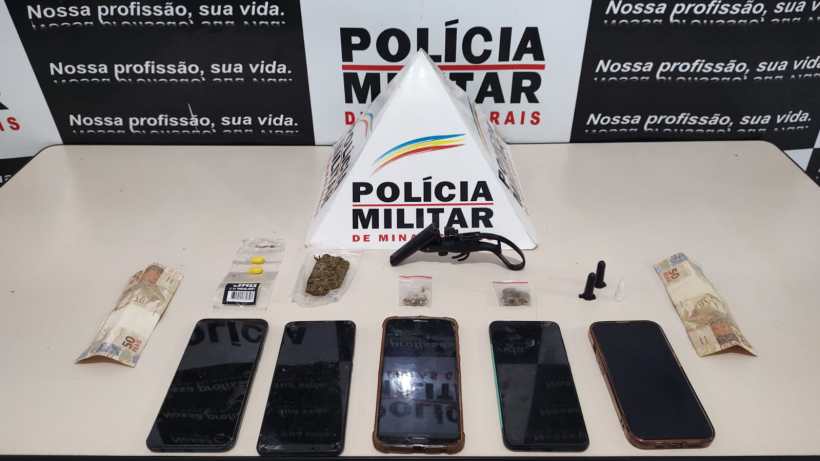 Material apreendido pela Polícia Militar, incluindo drogas, dinheiro e celulares, sobre uma mesa com fundo de banner da corporação.