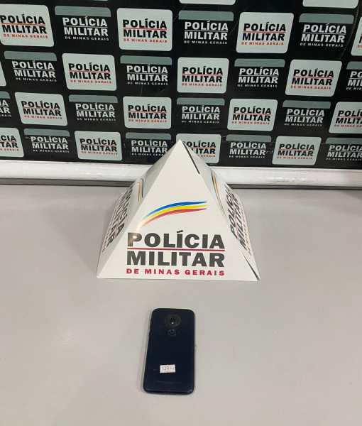 Um objeto em forma de pirâmide com o logo da Polícia Militar de Minas Gerais, colocado em uma mesa, ao lado de um celular.