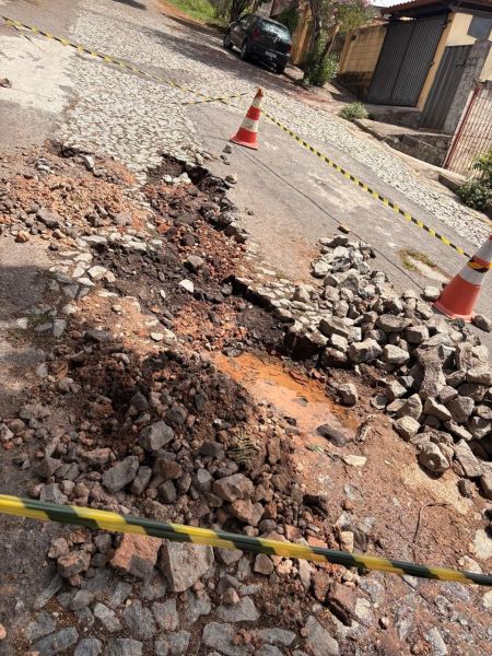 Buraco aberto na rua José de Alencar, em Divinópolis, com pedras soltas e sinais de intervenção da Copasa não concluída, cercado por cones e fitas para alertar os motoristas.
