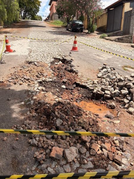 Buraco aberto em rua com cones e fitas de isolamento, resultado de obra inacabada da Copasa, comprometendo o trânsito e segurança em Divinópolis.