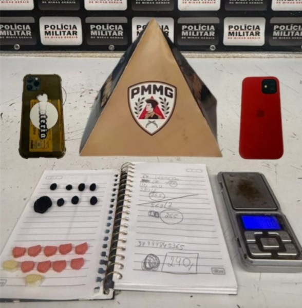 Objetos apreendidos durante operação policial, incluindo drogas, anotações, balança de precisão e celulares, em cima de uma mesa com fundo da Polícia Militar.