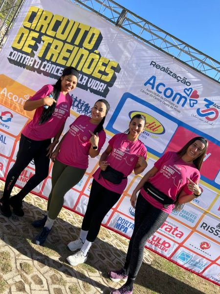 Quatro participantes do Circuito de Treinões, vestindo camisetas rosa, posam sorrindo em frente a um banner do evento, que promove a conscientização sobre o câncer.
