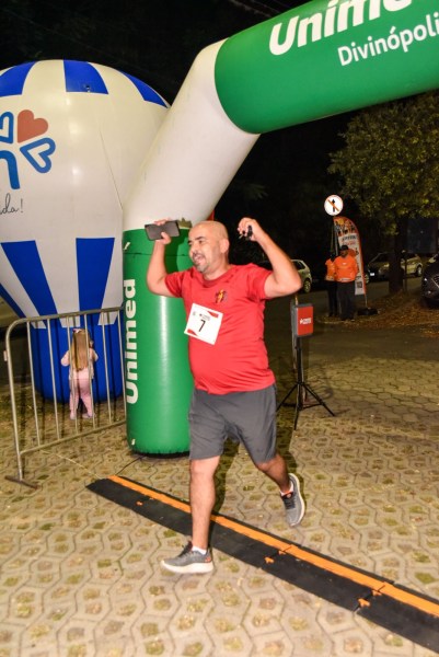 Homem correndo sob um arco de chegada do evento, vestindo camiseta vermelha e número de identificação, com um balão de ar quente ao fundo.
