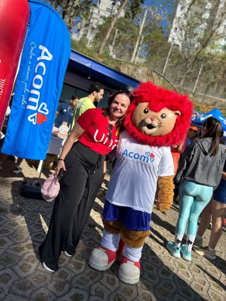 Uma mulher sorridente usando uma camiseta vermelha posa ao lado de um mascote leão, vestido com a camiseta da Acom, em um evento ao ar livre com barracas e árvores ao fundo.
