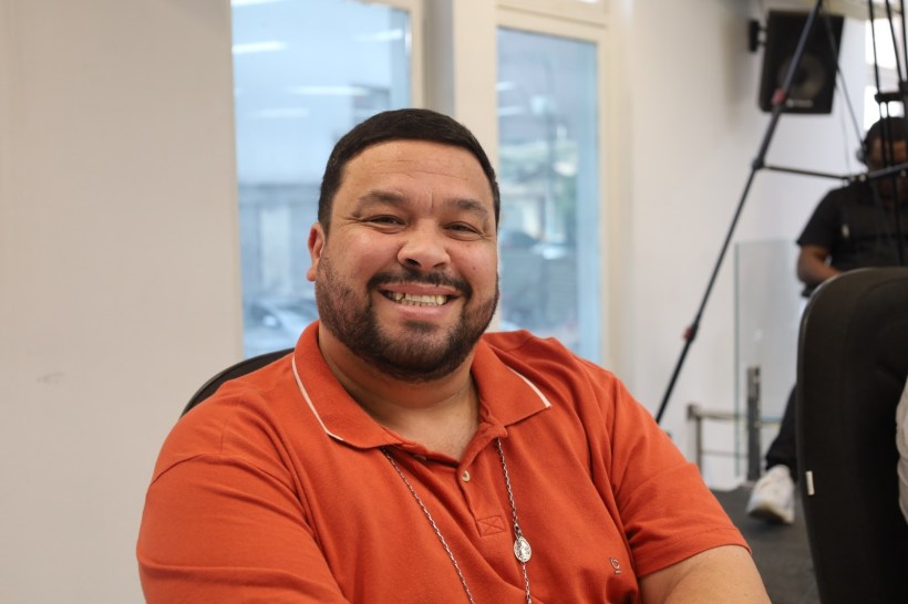 Retrato de um homem sorridente, com barba e cabelo curto, vestindo uma camiseta laranja. O fundo mostra um ambiente interno com janelas.
