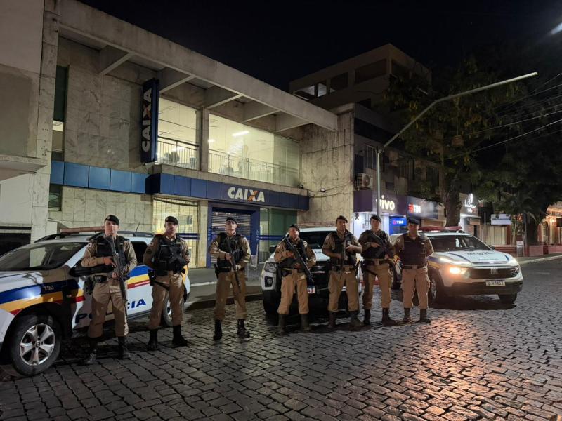 Sete policiais militares armados em frente a uma agência da Caixa Econômica Federal durante a madrugada, com viaturas estacionadas ao fundo em uma rua pavimentada.