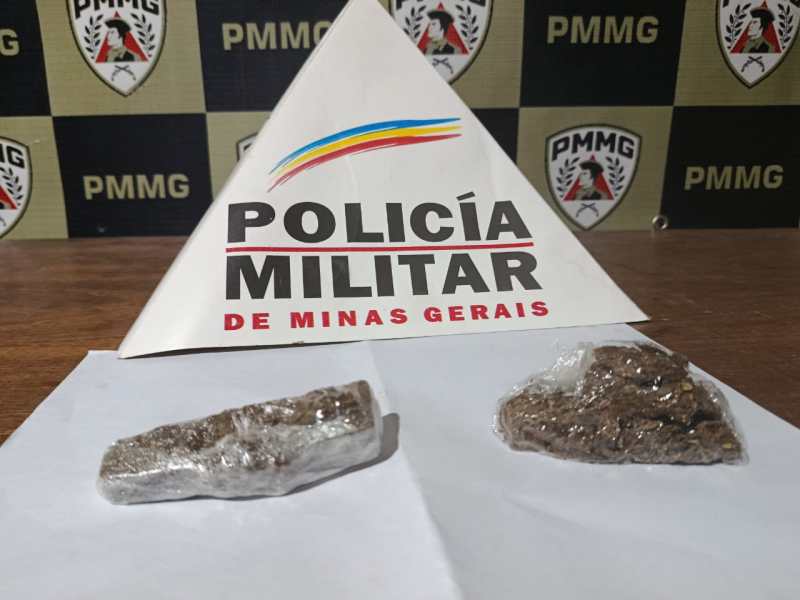 Dois pacotes de substância aparentemente ilegal, colocados em uma mesa com um cartaz da Polícia Militar de Minas Gerais ao fundo.