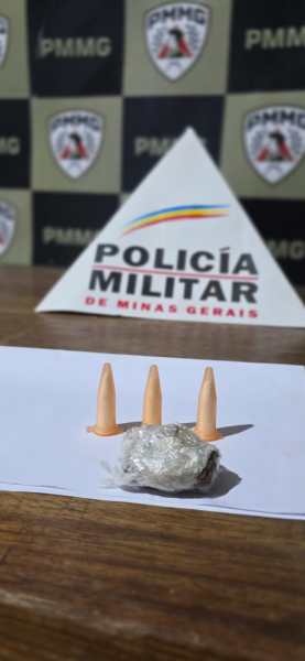 Tabela com um pedaço de substância aparentemente ilícita e três projéteis, com o fundo exibindo o logotipo da Polícia Militar de Minas Gerais.
