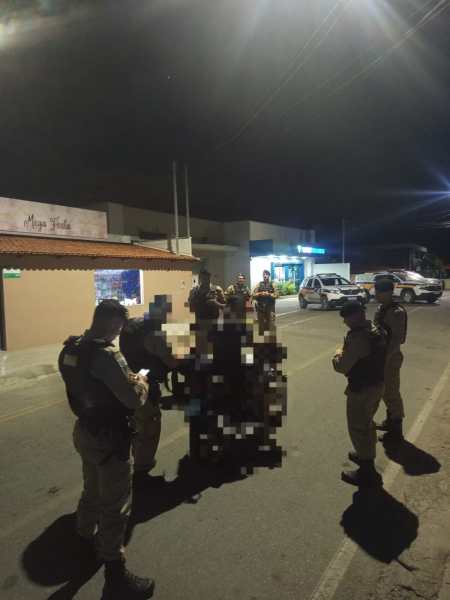 Policiais militares realizando uma abordagem durante uma operação noturna em uma rua, com veículos e uma loja visíveis ao fundo.