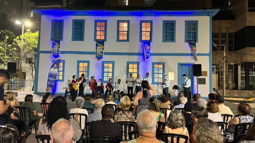 Ambiente do evento Seresta na Praça, com um grupo se apresentando em frente a uma casa colorida, iluminada em azul, enquanto o público aprecia a performance.
