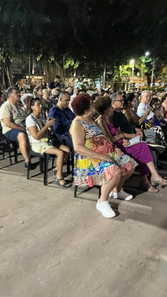 Público sentado assistindo ao evento Seresta na Praça, com várias pessoas de diferentes idades e estilos de roupas, em um espaço ao ar livre à noite.