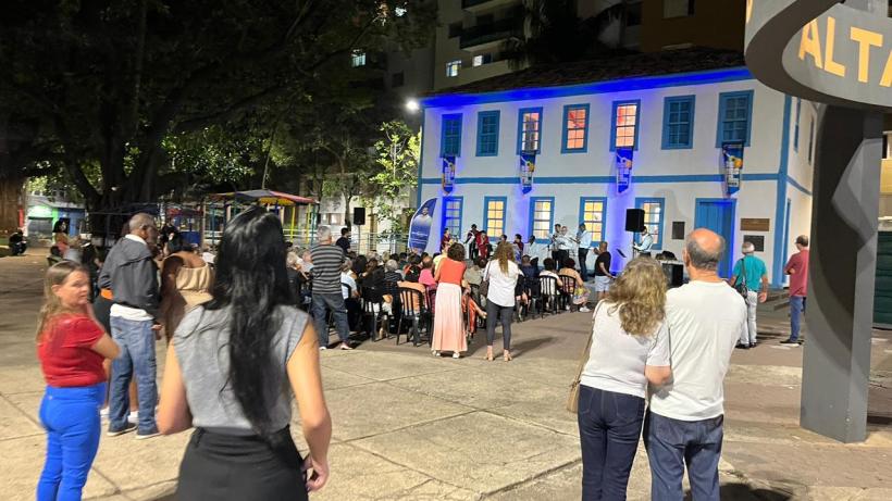 Plateia assistindo ao evento Seresta na Praça em Divinópolis, com um grupo de músicos se apresentando em um prédio histórico iluminado.