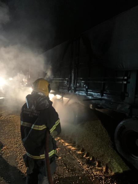 Bombeiro combater incêndio em carreta de transporte de grãos, com fumaça ao redor e parte da carga de milho espalhada no chão.