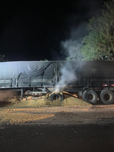 Carreta de transporte de grãos envolvida em incêndio, com fumaça saindo da roda e milho espalhado na pista.