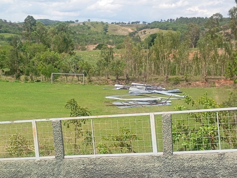 Vista de um campo de futebol com telhas metálicas danificadas e jogadas no chão, refletindo os danos causados por um vendaval, com vegetação ao fundo.