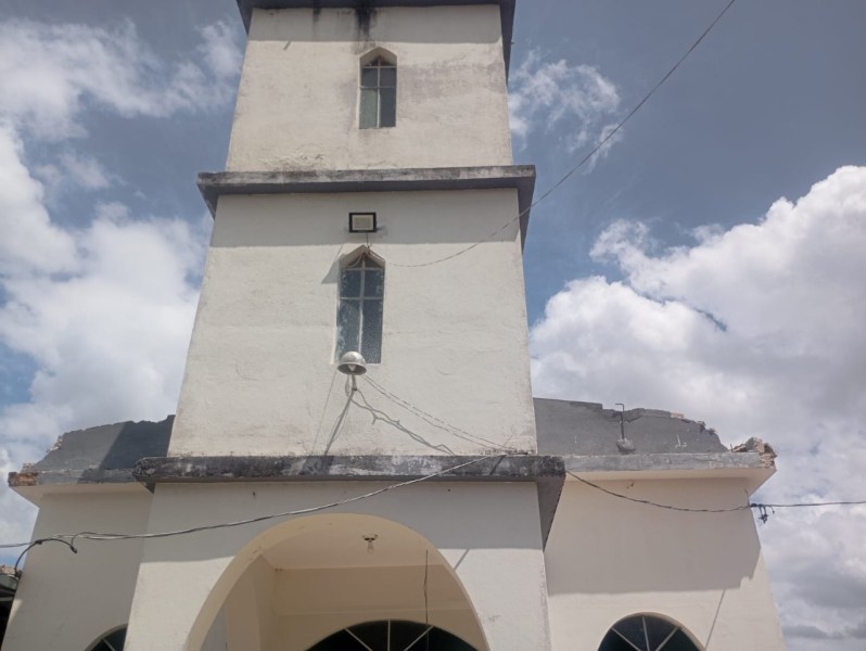 Vista da parte frontal de uma igreja com danos estruturais visíveis, incluindo a cobertura metálica parcialmente desabada devido a um vendaval, sob um céu parcialmente nublado.