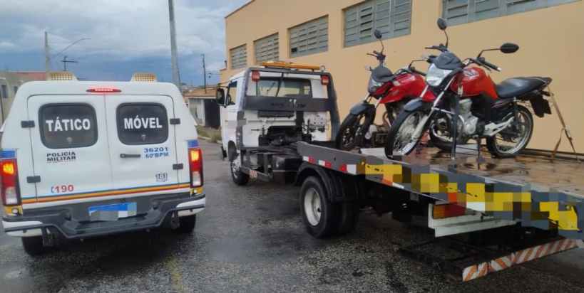Veículo da Polícia Militar e uma caminhonete de guincho com motocicletas sendo apreendidas em um pátio.