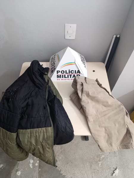 Uma mesa com uma jaqueta preta e verde, uma bermuda clara e um cone da Polícia Militar ao fundo.