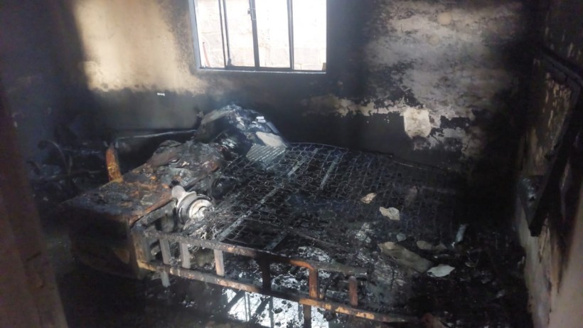 Interior de uma residência que foi parcialmente destruída por um incêndio, mostrando um espaço escurecido e danificado, com roupas e móveis queimados e uma cama em estado comprometido.